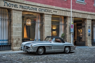A classic silver convertible car is parked on a cobblestone street in front of a building with a stone facade. The building, possibly a hotel or historical site, has large windows secured with iron bars and is illuminated from within by warm lighting. The sign above reads 'Hotel Pamplona Catedral'.