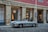 A classic silver convertible car is parked on a cobblestone street in front of a building with a stone facade. The building, possibly a hotel or historical site, has large windows secured with iron bars and is illuminated from within by warm lighting. The sign above reads 'Hotel Pamplona Catedral'.