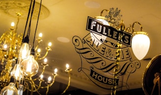 A decorative glass window displays the logo for Fuller's Ale & Pie House with elegant typography. Behind the glass, there are several light bulbs hanging from the ceiling, along with traditional chandeliers emitting a warm glow.