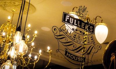 A decorative glass window displays the logo for Fuller's Ale & Pie House with elegant typography. Behind the glass, there are several light bulbs hanging from the ceiling, along with traditional chandeliers emitting a warm glow.