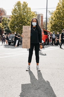 A person wearing a mask holds a protest sign in a street setting. The sign reads 'THEY WANT OUR RHYTHM BUT NOT OUR BLUES.' Other people are gathered in the background, some wearing masks, with trees and buildings visible.