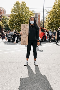 A person wearing a mask holds a protest sign in a street setting. The sign reads 'THEY WANT OUR RHYTHM BUT NOT OUR BLUES.' Other people are gathered in the background, some wearing masks, with trees and buildings visible.