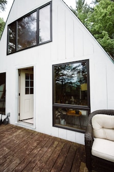 The image depicts the exterior of an A-frame house with white wooden paneling. There are large windows with black frames and reflections of trees visible in the glass. An open door leads inside the house, and on the porch, there is a wicker chair with a white cushion.