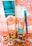 A collection of beauty products and accessories arranged on a decorative surface. The items include a tall metallic gold and turquoise bottle, a smaller matching bottle labeled as nail lacquer, a turquoise eyeliner pencil, and a set of large, circular, golden earrings. The background has an ornate, floral pattern in shades of peach and white.