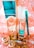 A collection of beauty products and accessories arranged on a decorative surface. The items include a tall metallic gold and turquoise bottle, a smaller matching bottle labeled as nail lacquer, a turquoise eyeliner pencil, and a set of large, circular, golden earrings. The background has an ornate, floral pattern in shades of peach and white.