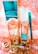 A collection of beauty products and accessories arranged on a decorative surface. The items include a tall metallic gold and turquoise bottle, a smaller matching bottle labeled as nail lacquer, a turquoise eyeliner pencil, and a set of large, circular, golden earrings. The background has an ornate, floral pattern in shades of peach and white.
