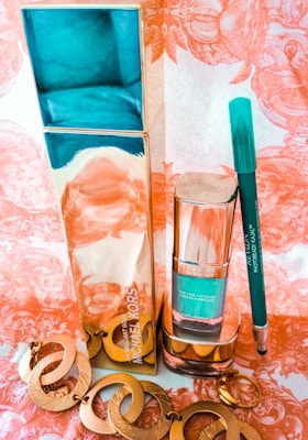 A collection of beauty products and accessories arranged on a decorative surface. The items include a tall metallic gold and turquoise bottle, a smaller matching bottle labeled as nail lacquer, a turquoise eyeliner pencil, and a set of large, circular, golden earrings. The background has an ornate, floral pattern in shades of peach and white.