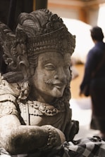 A detailed stone sculpture of a figure adorned with an intricate headdress and ornate garment, situated in an outdoor setting. The focus is on the craftsmanship of the sculpture with attention to facial features and decorative elements. In the background, there's a blurred silhouette of a person adding depth to the scene.