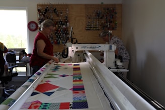 Two women are operating a large quilting machine in a well-lit room. The quilting machine has a colorful fabric with various geometric patterns on it. There is a thread rack against the wall, holding numerous spools of thread. A window to the left lets in natural light, and there's a clock nearby.