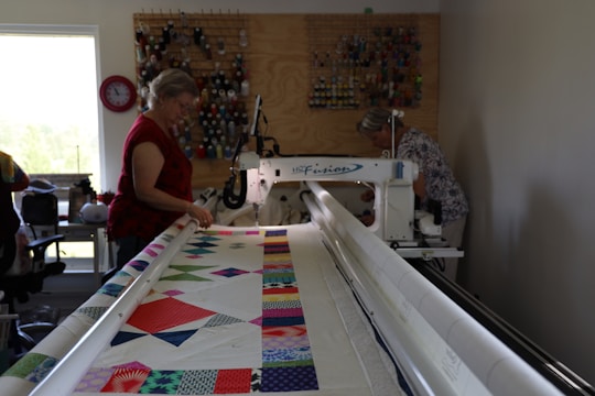 Two women are operating a large quilting machine in a well-lit room. The quilting machine has a colorful fabric with various geometric patterns on it. There is a thread rack against the wall, holding numerous spools of thread. A window to the left lets in natural light, and there's a clock nearby.