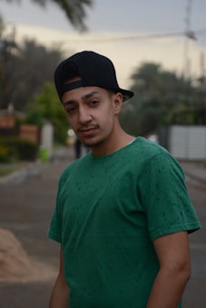 A young person is wearing a green t-shirt and black cap in an outdoor setting. The background shows a blurred view of trees and a street.