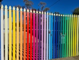 A picket fence painted in vertical stripes of bright colors ranging from yellow, orange, red, purple, blue, green to a light tan. The colors follow a rainbow gradient. Beyond the fence, there are some buildings and trees with a clear blue sky.