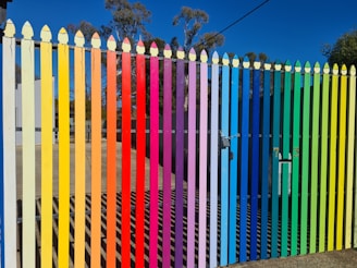 A picket fence painted in vertical stripes of bright colors ranging from yellow, orange, red, purple, blue, green to a light tan. The colors follow a rainbow gradient. Beyond the fence, there are some buildings and trees with a clear blue sky.