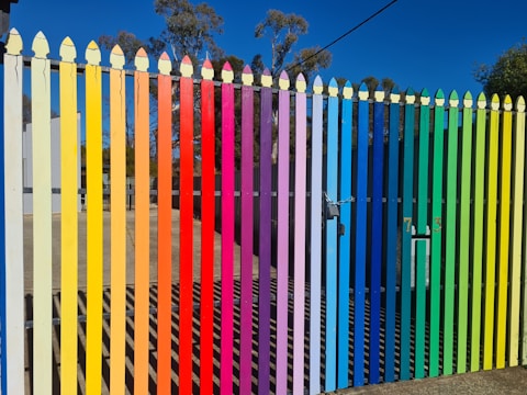 A picket fence painted in vertical stripes of bright colors ranging from yellow, orange, red, purple, blue, green to a light tan. The colors follow a rainbow gradient. Beyond the fence, there are some buildings and trees with a clear blue sky.