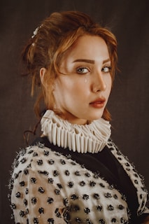 A woman with red hair is wearing a garment adorned with dark, textured embellishments and a white ruffled collar. Her expression is serious with a single tear visible on her left cheek against a neutral background.