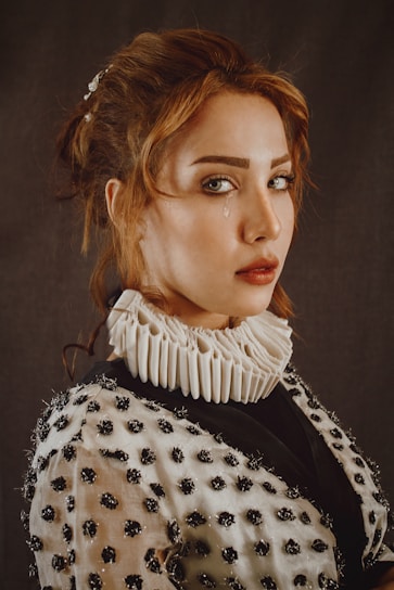 A woman with red hair is wearing a garment adorned with dark, textured embellishments and a white ruffled collar. Her expression is serious with a single tear visible on her left cheek against a neutral background.