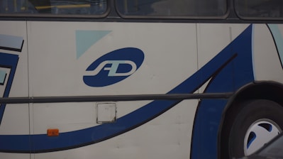 A close-up of the side of a bus displaying a logo with the letters 'SD'. The bus has a white exterior with blue and black curved lines as part of its design. The area shown includes a wheel and an orange reflector light.