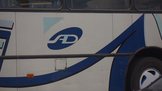 A close-up of the side of a bus displaying a logo with the letters 'SD'. The bus has a white exterior with blue and black curved lines as part of its design. The area shown includes a wheel and an orange reflector light.