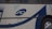 A close-up of the side of a bus displaying a logo with the letters 'SD'. The bus has a white exterior with blue and black curved lines as part of its design. The area shown includes a wheel and an orange reflector light.