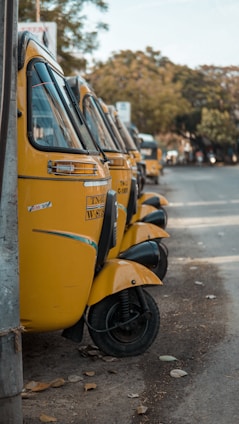 A row of yellow auto rickshaws is parked along the side of a road, with each vehicle closely lined up. The scene is set in an urban environment with trees and a road visible in the background. The rickshaws have distinct black wheels and a license plate visible on the front.