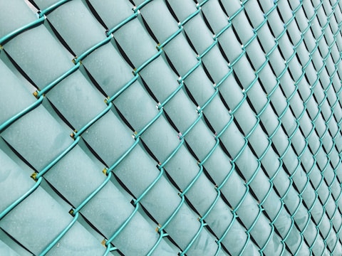 A close-up view of a wire fence with turquoise metal mesh, creating a repetitive, diamond-shaped pattern. The background appears to be a soft, light blue, possibly due to lighting or the material behind the fence.