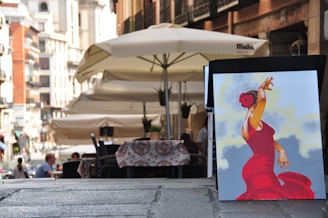 A quaint street scene with outdoor seating under large umbrellas. In the foreground, there's a vibrant painting of a flamenco dancer in a red dress with a red flower in her hair, set against a backdrop of soft blue and yellow hues.