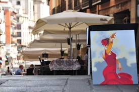 A quaint street scene with outdoor seating under large umbrellas. In the foreground, there's a vibrant painting of a flamenco dancer in a red dress with a red flower in her hair, set against a backdrop of soft blue and yellow hues.
