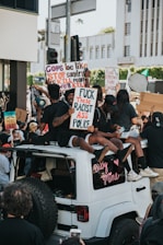 A group of people are gathered in a protest, with some individuals sitting on the roof of a white vehicle. They are holding up various signs in support of racial justice and police reform. The scene is dynamic and captures the energy of a large crowd engaged in activism.