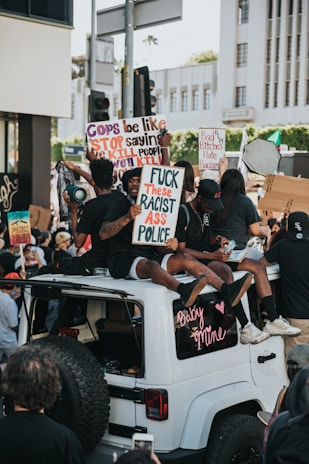 A group of people are gathered in a protest, with some individuals sitting on the roof of a white vehicle. They are holding up various signs in support of racial justice and police reform. The scene is dynamic and captures the energy of a large crowd engaged in activism.