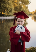 A young child wearing a red graduation cap and gown is smiling while holding a stuffed panda toy. The background features a serene, blurred nature scene with water and greenery.