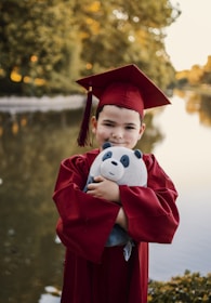 A young child wearing a red graduation cap and gown is smiling while holding a stuffed panda toy. The background features a serene, blurred nature scene with water and greenery.