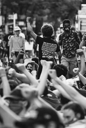 A group of people gather during a protest, many with their arms raised in a show of solidarity and unity. The central figure, with curly hair and wearing a t-shirt with text, stands with a raised fist. Others in the crowd are wearing masks, indicating a contemporary context.