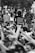 A group of people gather during a protest, many with their arms raised in a show of solidarity and unity. The central figure, with curly hair and wearing a t-shirt with text, stands with a raised fist. Others in the crowd are wearing masks, indicating a contemporary context.