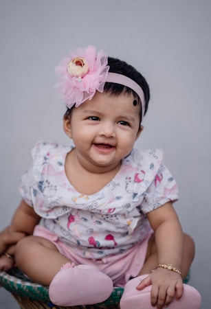 A joyful baby is sitting, dressed in a patterned outfit with pink elements. The baby wears a pink floral headband and light pink shoes, and has a delightful smile.