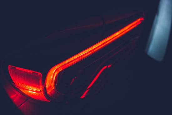 A close-up view of a car's rear brake light illuminated in a dark setting, with a sleek and modern design featuring sharp lines and glowing red hues.