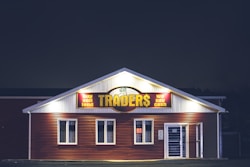 A brightly lit building at night with a sign reading 'TRADER$' and accompanying phrases like 'buy sell lend' and 'we pay cash'. The structure has a red and white facade and is adorned with several windows. A neon 'open' sign is visible in one of the windows.