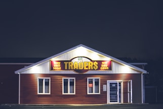 A brightly lit building at night with a sign reading 'TRADER$' and accompanying phrases like 'buy sell lend' and 'we pay cash'. The structure has a red and white facade and is adorned with several windows. A neon 'open' sign is visible in one of the windows.