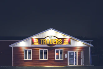 A brightly lit building at night with a sign reading 'TRADER$' and accompanying phrases like 'buy sell lend' and 'we pay cash'. The structure has a red and white facade and is adorned with several windows. A neon 'open' sign is visible in one of the windows.