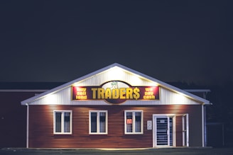 A brightly lit building at night with a sign reading 'TRADER$' and accompanying phrases like 'buy sell lend' and 'we pay cash'. The structure has a red and white facade and is adorned with several windows. A neon 'open' sign is visible in one of the windows.