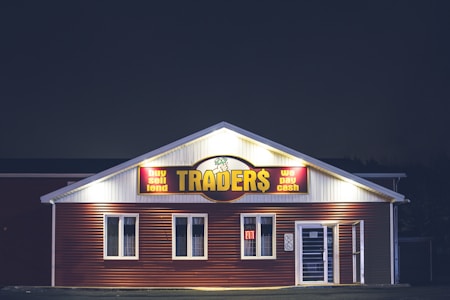 A brightly lit building at night with a sign reading 'TRADER$' and accompanying phrases like 'buy sell lend' and 'we pay cash'. The structure has a red and white facade and is adorned with several windows. A neon 'open' sign is visible in one of the windows.