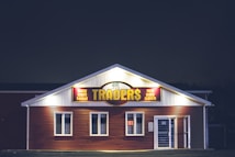 A brightly lit building at night with a sign reading 'TRADER$' and accompanying phrases like 'buy sell lend' and 'we pay cash'. The structure has a red and white facade and is adorned with several windows. A neon 'open' sign is visible in one of the windows.