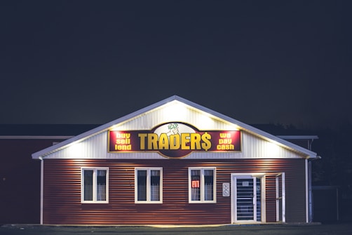 A brightly lit building at night with a sign reading 'TRADER$' and accompanying phrases like 'buy sell lend' and 'we pay cash'. The structure has a red and white facade and is adorned with several windows. A neon 'open' sign is visible in one of the windows.