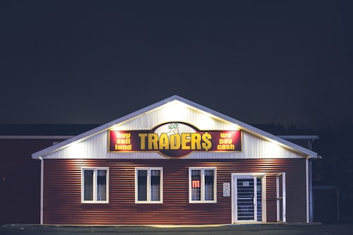 A brightly lit building at night with a sign reading 'TRADER$' and accompanying phrases like 'buy sell lend' and 'we pay cash'. The structure has a red and white facade and is adorned with several windows. A neon 'open' sign is visible in one of the windows.