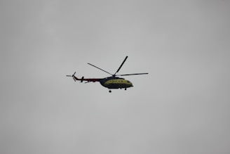 A helicopter with a green and red color scheme is flying against a cloudy gray sky. The helicopter is viewed from the side, and its rotor blades are clearly visible.
