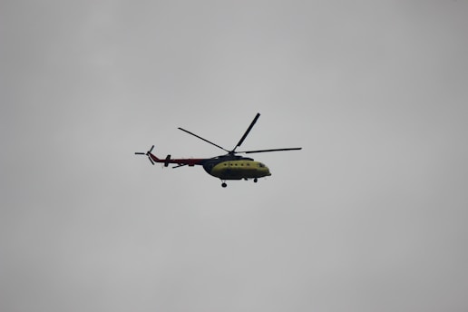 A helicopter with a green and red color scheme is flying against a cloudy gray sky. The helicopter is viewed from the side, and its rotor blades are clearly visible.
