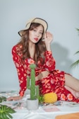 A young woman with wavy brown hair is seated, wearing a stylish floral red dress and a wide-brimmed hat. The setting includes a few green plants, a pineapple, and some scattered papers or magazines on a white surface.
