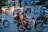 Three people, including a child, are riding a motorbike in a busy city street. All of them are wearing helmets. They are surrounded by other motorbikes also being ridden by people who are wearing helmets. The background displays an urban scene with illuminated buildings and blurred lights.