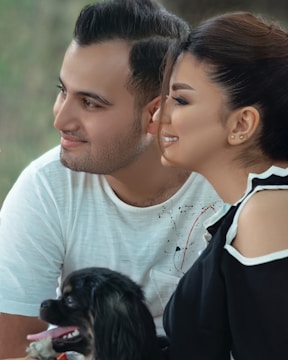 A couple is seated closely together, smiling and looking towards the left. They are accompanied by a small black and white dog that appears cheerful. The background is a soft blur of green and brown tones, suggesting an outdoor setting.