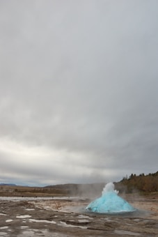 A geyser is in the process of erupting, with a vivid blue bubble of water and steam rising above the ground. The surrounding landscape is rocky and arid, and the overcast sky adds to the dramatic scene.