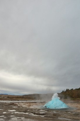 A geyser is in the process of erupting, with a vivid blue bubble of water and steam rising above the ground. The surrounding landscape is rocky and arid, and the overcast sky adds to the dramatic scene.